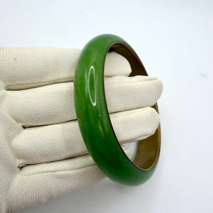 May include: A smooth, rounded bangle bracelet featuring a vibrant, solid green outer surface and a contrasting metallic inner band. The design is simple and circular, showcasing a polished finish that reflects light across its curved exterior.