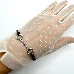 May include: A sheer off-white glove featuring a delicate pattern of small, light-colored dots. A brown cord bracelet with a silver-toned metal horse bit connector is wrapped around the wrist area of the glove, secured with simple knots.