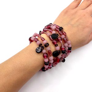 May include: A multi-strand wrap bracelet featuring a variety of beads in shades of deep burgundy, translucent red, and matte light pink. The design includes round beads, faceted glass beads, and two distinct dark teardrop-shaped charms.