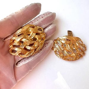 May include: A pair of gold-coloured clip-on earrings. Each earring has a woven, basket-style design. The earrings are shown on a pink surface. The earrings have a shiny, metallic finish.