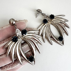 Op de afbeelding: Zilverkleurige statement oorbellen met een gestileerd bladontwerp. Elke oorbel heeft een cluster van donkerblauwe en heldere edelstenen. De oorbellen hebben een stekersluiting en een gepolijste afwerking, geschikt voor formele gelegenheden.