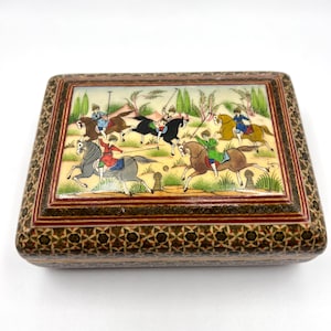 May include: A rectangular wooden box with a painted lid depicting riders on horseback. The box has a decorative border with a geometric pattern. The scene shows figures in colourful attire, engaged in a sport or competition. The box is likely for storing small items.