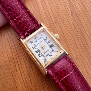 Könnte beinhalten: Eine rechteckige, goldfarbene Quarzuhr mit weißem Zifferblatt und schwarzen römischen Ziffern. Die Uhr hat ein rotes, strukturiertes Lederarmband. Auf dem Zifferblatt steht "PATRICK ARNOLD" und "QUARTZ".