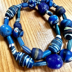 以下が含まれることがあります： さまざまな青いガラスビーズを使用した手作りのネックレス。ビーズには、無地の青い球体、ストライプのシリンダー、白と青の模様が施されたユニークな形のビーズが含まれます。ネックレスは青いコードに通されており、金属製の留め金が付いています。