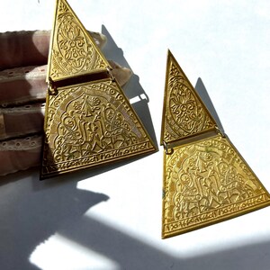 May include: A pair of gold-coloured, triangular vintage earrings. Each earring has detailed embossed patterns of floral and figurative designs. The earrings have a hinged clasp and are displayed on a white surface.