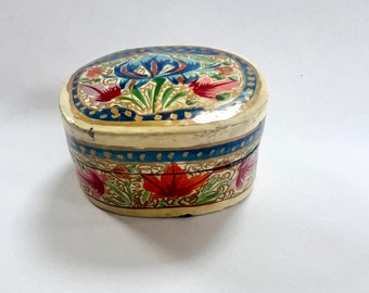 Pode incluir: Uma pequena caixa de joias oval com um design floral. A caixa é pintada com uma variedade de cores, incluindo vermelho, azul, verde e dourado. A tampa da caixa apresenta uma grande flor azul com um centro amarelo.
