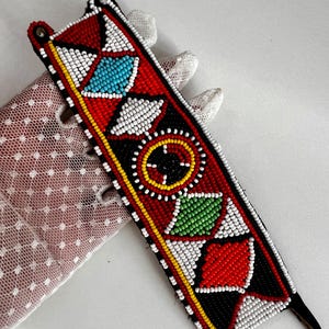 May include: A beaded choker necklace with a geometric design. The necklace features red, white, black, yellow, blue, and green beads. The choker has a black backing and brown leather ties.