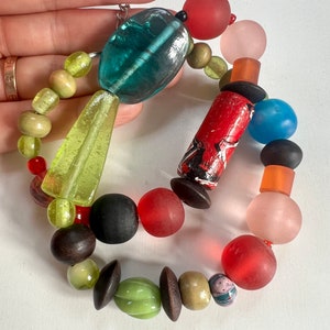 May include: A vibrant beaded bracelet with diverse shapes and materials. It features green, red, black, and pink beads, alongside wooden and glass components. The design combines geometric and organic forms, creating a unique accessory. The bracelet is approximately 20 cm in circumference.