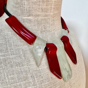 Puede incluir: Un collar llamativo con elementos de vidrio rojo y transparente alternados. El collar tiene una banda negra curvada y se muestra en un maniquí. Las piezas de vidrio son geométricas y tienen un acabado brillante.