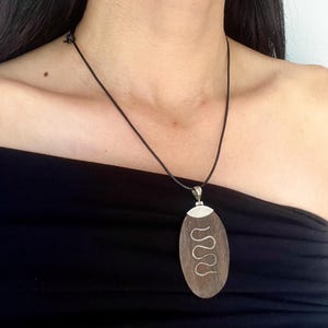 Puede incluir: Collar con colgante de madera con diseño de serpiente plateada. El colgante ovalado cuelga de un cordón negro. El collar se lleva sobre una camiseta negra.