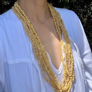 May include: A multi-strand necklace composed of small, light-yellow beads. The necklace is draped over a white shirt, creating a contrast in color. The beads appear to be natural, possibly shell or seed-based, and are densely packed.