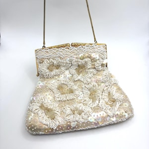 May include: A cream-coloured handbag featuring intricate white beadwork and iridescent sequins arranged in floral-like patterns. The bag has a gold-toned metal frame with an ornate clasp and a thin, matching gold-toned metal chain shoulder strap.