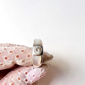 May include: A silver ring with a rectangular design. The ring features a central, round, clear gemstone and two rectangular mother-of-pearl inlays. The ring is held by a pink lace glove.