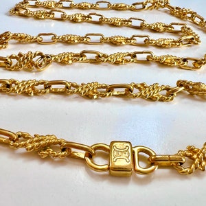Puede incluir: Un collar dorado con una combinación de eslabones tipo cuerda y rectangulares. El collar presenta un cierre con una pieza central rectangular con un logotipo. La joya se exhibe sobre una superficie blanca.