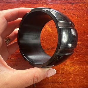 May include: A black, wide bangle bracelet with a glossy finish. The bracelet has a series of raised, rectangular designs around the band. The bracelet is held by a hand, showcasing its size and design. The bracelet is a fashion accessory.