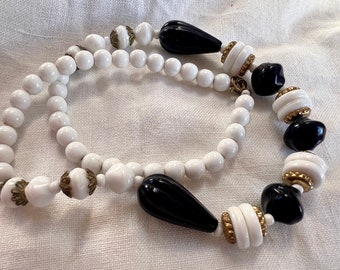 Op de afbeelding: Een witte en zwarte kralenketting met gouden accenten. De ketting is gemaakt van ronde witte kralen en zwarte druppelvormige kralen.