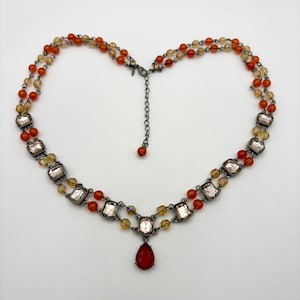 Op de afbeelding: Een meerstrengs ketting met oranje en gele kralen, rechthoekige heldere kristallen in donkere metalen zettingen en een enkele druppelvormige rode hanger in het midden, voorzien van een donkere metalen ketting met een verstelbare sluiting.