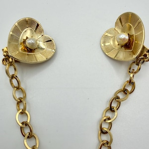 May include: A gold-tone metal cardigan clip featuring two heart-shaped clasps with a radial sunburst texture. Each heart is centred with a small white faux pearl. The two clasps are connected by a gold-tone oval link chain.
