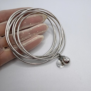 May include: A set of four thin, silver-toned metal bangle bracelets. The bangles are connected by a small silver ring holding a round, polished silver bead charm and a small, flat, circular silver tag engraved with the word "mink".
