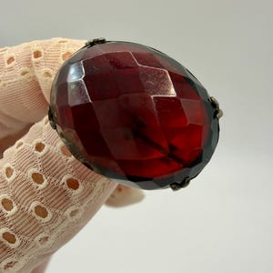 May include: A large, faceted, oval-shaped, dark red gemstone set in a dark metal frame. The gemstone has a complex, geometric cut, reflecting light. The setting appears to be antique or vintage, with a detailed design.