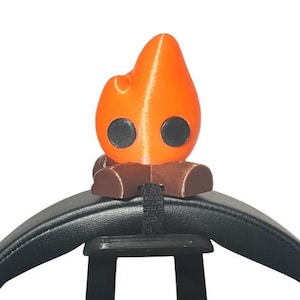 Op de afbeelding: Een 3D-geprint oranje vlamvormig accessoire met zwarte ogen en een bruine stamvoet, bevestigd aan een zwart object met een zwarte band. De vlam heeft een gladde textuur en staat tegen een witte achtergrond.