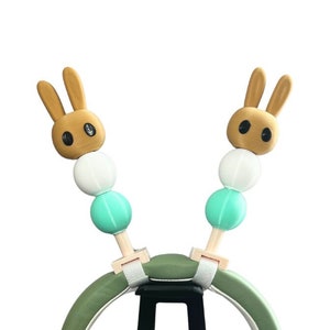 May include: Green headphones with white earcups and a black stand. The headphones have brown bunny ear-shaped charms with white and blue beads attached.
