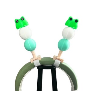 May include: Green headphones with frog-shaped charms on the headband. The headphones are on a black stand.