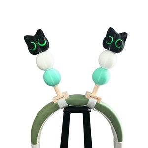 Puede incluir: Auriculares verdes con cubiertas de orejas de gato negras en un soporte negro. Las orejas de gato tienen ojos verdes y cuentas blancas y azul claro.