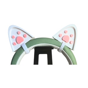 May include: White cat ear headband with pink paw prints on each ear. The headband is green and white and is attached to a black stand.