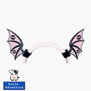 May include: A pair of pink and black bat wings with skull details attached to a white headband. The headband is made of a flexible material and has black velcro straps for attaching the wings.