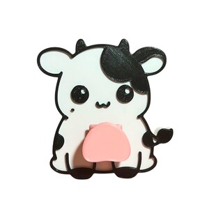 May include: A cute, cartoon-style cow figurine with a white body, black spots, and a pink udder. The cow has large, black eyes and a smiling mouth. The outline is black, and the figurine is set against a white background.