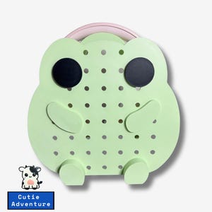 Op de afbeelding: Een groene kikker-vormig prikbord met zwarte ogen en een roze handvat. Het prikbord heeft een raster met gaten om kleine items aan te hangen.
