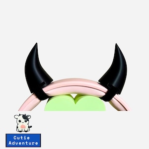 May include: Two black devil horns with a pink and green foam ring. The horns are attached to the ring with black straps. The text "Cutie Adventure" is visible in the image.