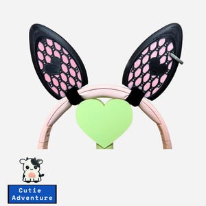 May include: A pink headband with black and pink bunny ears. The ears have a heart-shaped design. A green heart is centered on the headband. The text "Cutie Adventure" is below a cartoon cow.