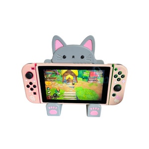 May include: A pink and gray cat-shaped stand for a Nintendo Switch console. The stand has a gray cat face with pink ears and paws. The Switch console is in a pink and gray case with a game screen showing a video game.