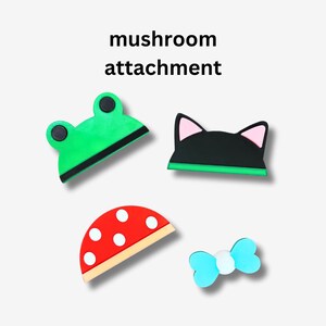Op de afbeelding: Vier kleurrijke paddenstoelbevestigingen voor een schoonmaakgereedschap. De bevestigingen zijn gevormd als een kikker, een kat, een paddenstoel en een strik. De tekst "mushroom attachment" staat boven de bevestigingen.
