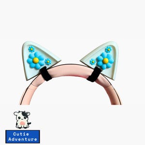 May include: A pair of light blue cat ear headphones with a pink headband. The cat ears have blue flowers with yellow centres.