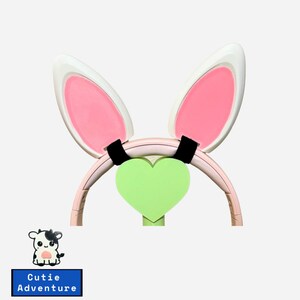 May include: Pink bunny ear headband with a green heart charm. The headband is made of plastic and has a soft, comfortable feel. The bunny ears are large and fluffy, and the heart charm is attached to the headband with a black elastic band.