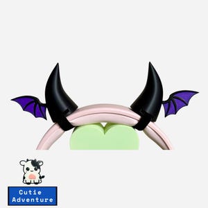 May include: A pair of black devil horns with purple wings attached to a pink headband. The headband is wrapped around two heart-shaped cushions.