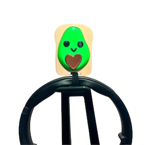 May include: A green avocado with a brown heart on a beige toast-shaped headphone stand. The avocado has a smiling face with black eyes.