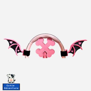 May include: A pink and black bat wing headband with a pink cross-shaped design in the center. The wings are black with pink heart accents. The headband has a light pink padded band and black velcro straps.