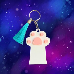 May include: White paw print keychain with a pink paw pad and a turquoise tassel. The keychain is against a purple and blue galaxy background.