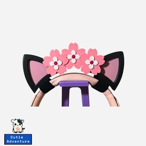 May include: A cat ear headband with pink inner ears and black trim, adorned with pink flower accents. The headband is supported by a purple stand. The text "Cutie Adventure" is displayed on a blue rectangle.
