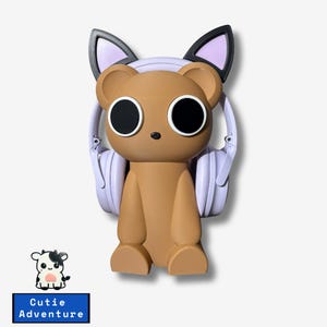May include: A brown bear-shaped figurine with large black eyes and a small black nose. The bear has light purple headphones around its neck and cat ears. The text "Cutie Adventure" is on a blue rectangle below a cow illustration.
