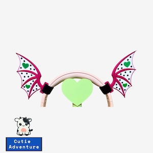 May include: Pink and white polka dot devil wings with green hearts attached to a light brown wooden hoop. The wings are made of a soft, flexible material and are perfect for adding a touch of whimsy to any outfit.