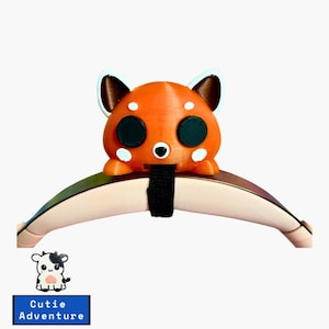 May include: A red panda-shaped headphone charm with black eyes and white spots. The charm is attached to a black strap and is sitting on a pink headband.