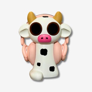 May include: A white cow-shaped speaker with black spots and pink ears. The speaker has large black eyes and a pink nose. It is wearing pink headphones.