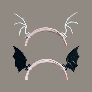 May include: Two pairs of headphone clips, one white and one black. The white clips are shaped like skeletal wings, while the black clips are shaped like bat wings.