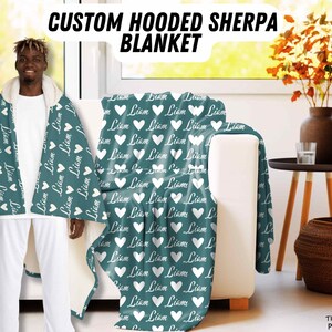 K&ouml;nnte beinhalten: Eine tealfarbene Kapuzen-Sherpa-Decke mit wei&szlig;en Herzen und dem Namen "Liam" darauf gedruckt. Die Decke wird von einer Person getragen.