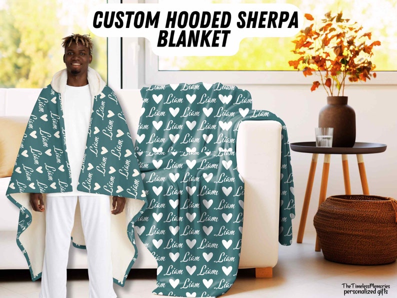 K&ouml;nnte beinhalten: Eine tealfarbene Kapuzen-Sherpa-Decke mit wei&szlig;en Herzen und dem Namen "Liam" darauf gedruckt. Die Decke wird von einer Person getragen.